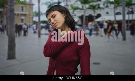 La giovane donna ispanica in maglione Bordeaux afferra e tiene le spalle per il dolore mentre si trova in strada; disagio. Foto Stock