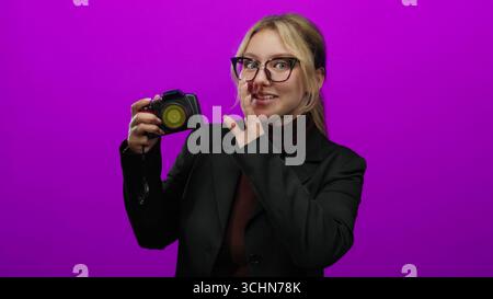La giovane donna bionda che tiene la macchina fotografica sussurra umoristicamente il segreto su un vivace sfondo rosa in un ambiente isolato, mostrando un'espressione giocosa Foto Stock