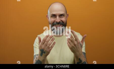 Gesti di uomo dalla barba calva con le mani contro la parete arancione, che mostrano tatuaggi ed espressioni invitanti, indossando una camicia gialla in un set da studio isolato Foto Stock