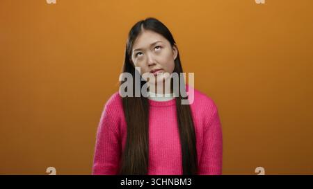 Donna con i capelli lunghi in un maglione rosa in piedi su uno sfondo arancione con un'espressione contemplativa su una parete isolata. Foto Stock
