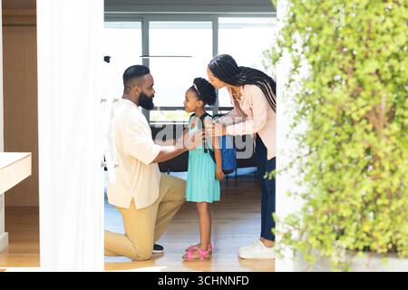 I genitori aiutano la figlia con lo zaino, si preparano per la scuola a casa, sorridono calorosamente Foto Stock