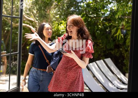 Due giovani donne, una con lunghi capelli marroni in abito rosso, entrano felicemente nel patio illuminato dal sole Foto Stock