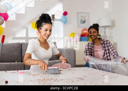 Ripulisce dopo la festa, due amiche diverse che ripuliscono il soggiorno con sorrisi, a casa Foto Stock