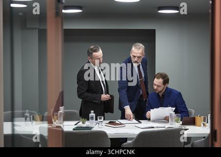 Tre uomini caucasici, due maturi e uno giovane, collaborano in una moderna sala conferenze, discutono di documenti e analizzano i dati finanziari nei documenti durante la riunione di lavoro Foto Stock