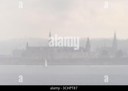 Castello di Kronborg attraverso Mist Helsingør Danimarca / Helsingør, Danimarca — il castello di Kronborg, patrimonio dell'umanità dell'UNESCO, è parzialmente oscurato dalla nebbia sulla costa di Helsingør. Lo storico castello, situato sulla punta nord-orientale dell'isola di Zelanda, è visibile attraverso lo stretto di Øresund, con una barca a vela che naviga sull'acqua in primo piano e la sagoma delle torri e delle guglie del castello che emergono dalla foschia atmosferica. Il castello è rinomato per il suo ruolo di ambientazione per l'Amleto di Shakespeare e per la sua importanza strategica nel controllo del suono. Foto Stock