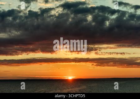 Alba sul Mar di Kattegat vicino a Strömstad Svezia // STRÖMSTAD, Svezia: Una spettacolare alba illumina il cielo sul Mar di Kattegat vicino a Strömstad, Svezia. Nuvole scure e pesanti sono intervallate da bande di arancione e giallo vibranti, che segnalano l'alba. Il sole, una sfera luminosa, sbircia sull'orizzonte lontano, che getta un caldo bagliore sull'acqua. Strömstad è una città costiera della provincia di Västra Götaland, nota per il suo arcipelago e la vicinanza alla Norvegia. Il Kattegat è uno stretto tra Danimarca e Svezia, che collega il Mar Baltico al Mare del Nord. Foto Stock