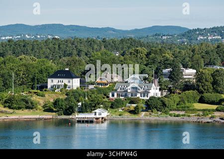 Case sul lungomare di Oslofjord Lysaker Norway // LYSAKER, Norway — le case costeggiano il lungomare dell'Oslofjord a Lysaker, Akershus. L'area residenziale e' annidata tra lussureggianti alberi verdi e colline ondulate. Lungo la costa sono visibili diverse grandi case dall'architettura distintiva. L'Oslofjord è una famosa area ricreativa e un canale navigabile chiave per la regione. Foto Stock