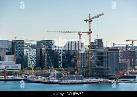 Gru da cantiere che si affacciano sugli edifici Waterfront Oslo Norvegia // OSLO, Norvegia — le gru da cantiere sono attive sugli edifici sul lungomare di Oslo, Norvegia, una città in fase di sviluppo urbano significativo. La scena cattura la trasformazione in corso dell'area portuale, con progetti architettonici moderni in corso. Diverse gru, tra cui spiccano le gru gialle a torre e una blu, dominano lo skyline, indicativo di una costruzione su larga scala. Tra le strutture visibili ci sono edifici di uffici contemporanei, alcuni caratterizzati da distintive facciate geometriche. Il lungomare stesso mostra chiatte e. Foto Stock