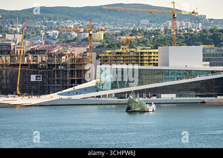 Si trova Sculpture Oslo Opera House Oslo Norway // OSLO, Norway — l'isola scultorea che giace di Monica Bonvicini si trova in acqua di fronte al Teatro dell'Opera di Oslo. Il Teatro dell'Opera, progettato da Snøhetta, è un importante punto di riferimento culturale a Oslo, Norvegia, noto per la sua architettura distintiva che consente ai visitatori di camminare sul suo tetto. Le gru da cantiere sono visibili sullo sfondo, a indicare lo sviluppo in corso nell'area. Il Teatro dell'Opera di Oslo è stato inaugurato ufficialmente nel 2008 e da allora è diventato un simbolo del moderno paesaggio architettonico della città. L'opera d'arte che mente è un annuncio notevole Foto Stock