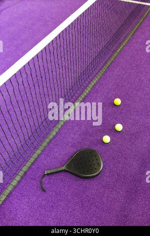 Racchetta di padel nera che si trova vicino a tre palle gialle sul campo viola, con rete che si estende diagonalmente Foto Stock
