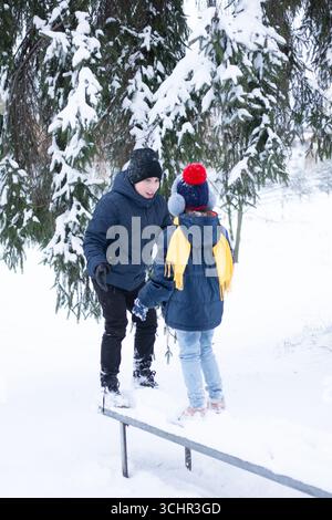 Un ragazzo e una ragazza, vestiti in modo caldo con cappelli, sciarpe e giacche, interagiscono giocosamente su una panchina innevata. La scena è ambientata in una foresta invernale innevata, W Foto Stock