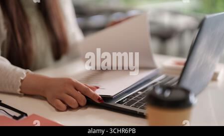 La mano di un adulto esamina importanti documenti aziendali sulla scrivania vicino a un computer portatile. Un professionista efficiente gestisce le attività in un ambiente di ufficio moderno. Foto Stock