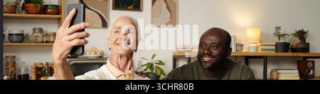 Donna caucasica anziana con smartphone che scatta selfie con il giovane uomo nero seduto accanto al sorriso, entrambi che guardano il dispositivo in un ambiente accogliente della cucina di casa Foto Stock
