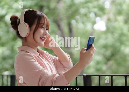 La giovane donna asiatica adulta gode di momenti sereni all'aperto con cuffie wireless e smartphone che abbracciano la libertà digitale della natura Foto Stock