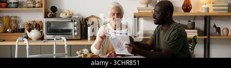 Donna caucasica anziana sorridente mentre si siede a tavola con un giovane nero che discute di documenti, camminatore visibile in primo piano, scaffali della cucina e elettrodomestici sullo sfondo Foto Stock
