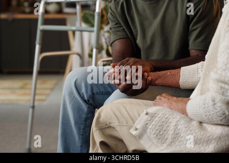 Giovane uomo adulto di colore che tiene per mano una donna caucasica anziana seduta insieme, mostrando sostegno e compassione, vista parziale focalizzata su mani e braccia, camminatore visibile sullo sfondo Foto Stock