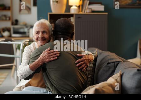Donna caucasica anziana sorridente mentre abbraccia l'uomo nero sul divano, entrambi seduti vicino in salotto, donna anziana che mostra affetto e calore durante l'abbraccio Foto Stock
