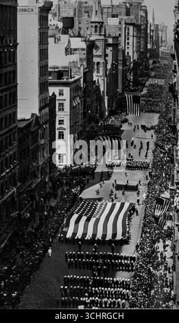 Fotografia di una parata patriottica sulla Fifth Avenue, New York City, che mostra una massiccia bandiera americana trasportata dal Carpet Club, pubblicata su l'Illustration, 17 giugno 1916. La scena mette in evidenza le manifestazioni pubbliche statunitensi di orgoglio nazionale durante la prima guerra mondiale, prima dell'ingresso formale dell'America nel 1917. Le folle fiancheggiano il viale mentre bande, soldati e gruppi civici marciano sotto i torreggianti edifici di Manhattan, simboleggiando l'unità americana e il crescente coinvolgimento negli affari mondiali. Foto Stock