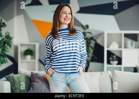 Giovane donna in felpa casual a righe sorridente in interni in un elegante soggiorno Foto Stock