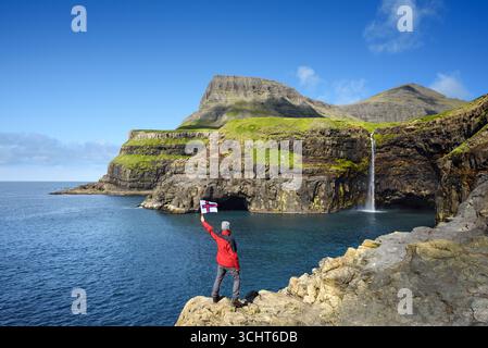 Uomo in giacca rossa che regge la bandiera delle Isole Faroe in piedi sulla scogliera godendo di una vista panoramica della cascata Mulafossur vicino a Gasadalur, Isole Faroe Foto Stock