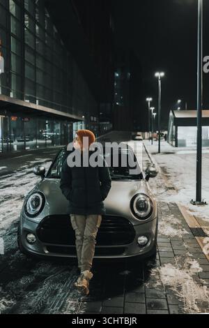 Donna con berretto arancione appoggiata su un'auto d'argento di notte in inverno, città innevata. La donna si riposa dopo un viaggio su strada. Foto Stock