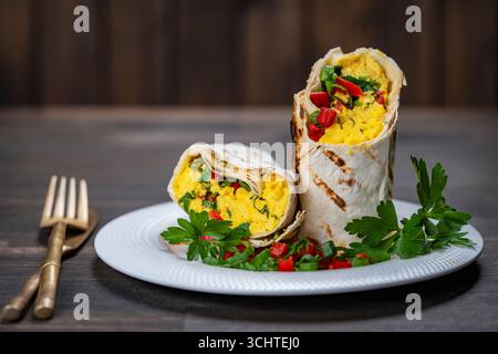 Colazione sana e alla moda fatta in casa uova burrito con verdure ed erbe aromatiche in un piatto bianco su un tavolo di legno, primo piano. Omelette all'uovo avvolta in una pita brea Foto Stock