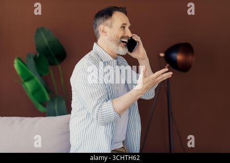 Uomo d'affari felice e maturo che parla al telefono con interni moderni in stile loft con impianti che lavorano da casa su un nuovo progetto Foto Stock