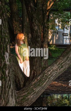 Una donna con camicetta verde e pantaloni bianchi si trova tra i tronchi di grandi alberi, illuminata dalla calda luce del tramonto Foto Stock