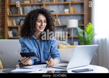 Giovane donna che lavora da casa, che calcola e prende appunti mentre utilizza il suo laptop per rivedere i rapporti finanziari, gestendo le finanze domestiche in modo efficiente e confortevole Foto Stock
