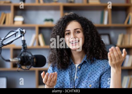 Giovane donna felice che registra un podcast da casa, parla al microfono e gesta con le mani, circondata da libri sugli scaffali nel suo accogliente studio domestico Foto Stock