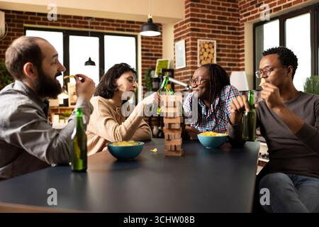 Gruppo multietnico di amici che si godono una pausa dal gioco, condividendo spuntini e bevande intorno al tavolo. Le donne caucasiche e afroamericane fanno il tifo con le bottiglie di birra mentre gli uomini sgranocchiano le patatine. Foto Stock