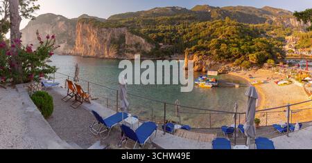 Spiaggia e scogliere costiere a Paleokastritsa Corfù in Grecia con lettini ombrelloni e barche al tramonto Foto Stock