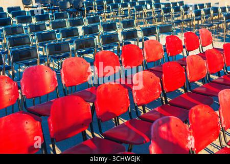 File di sedie vuote sono disposte all'aperto, con i sedili anteriori di colore rosso brillante e quelli dietro di loro di colore nero. La luce del sole proietta ombre vibranti Foto Stock