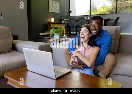 Coppie diverse che ridono insieme mentre guardano il laptop a casa, godendosi un momento di relax Foto Stock