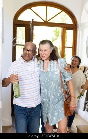 Coppia senior che arriva a casa, tenendo in mano il vino e sorridendo calorosamente con amici anziani diversi Foto Stock