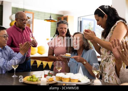 Amici anziani che festeggiano il compleanno a casa, donna che fa saltare la candela sulla torta Foto Stock