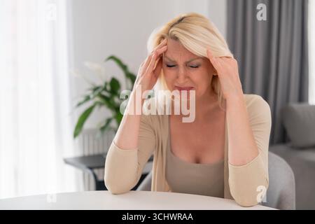 Donna bionda di mezza età seduta da sola al tavolo della cucina, coprendo il viso con le mani, piangendo con disperazione e tristezza, crollo emotivo a casa Foto Stock