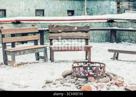 panche in legno intorno al focolare con pietre e ceneri nel giardino innevato, area ricreativa all'aperto rustica invernale concetto di campeggio, turismo e avventura Foto Stock
