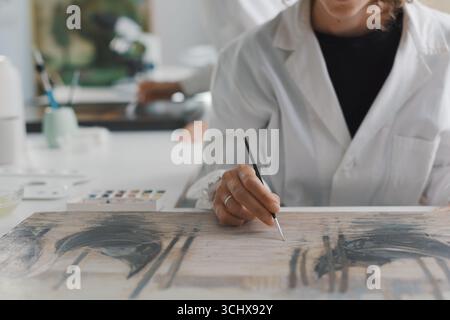 Conservatrice di pittura che ripristina un'opera d'arte usando un pennello, è seduta alla sua scrivania e lavora Foto Stock