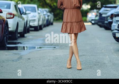 Una donna irriconoscibile cammina lungo una strada della città, indossando un abito marrone e tacchi alti. Il percorso è fiancheggiato da auto parcheggiate, che riflettono un'atmosfera vivace Foto Stock