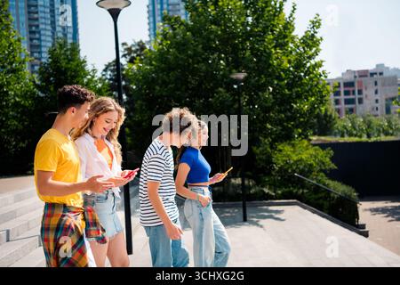 Gruppo di giovani diversi che si godono una giornata di sole all'aperto, camminano e utilizzano smartphone in un paesaggio urbano moderno Foto Stock