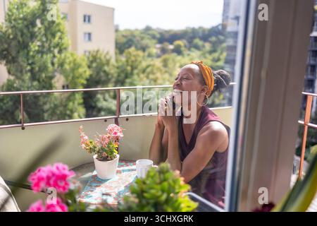 Donna che beve caffè e usa il telefono cellulare sul balcone di casa Foto Stock