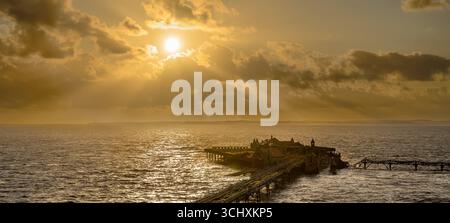 Il sole tramonta al vecchio molo di Birnbeck a Weston Super Mare, Somerset, Regno Unito, con i raggi del sole che scendono. Il molo è impostato per essere ripristinato. Foto Stock