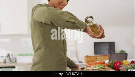 Donna che versa l'olio dal vasetto alle verdure sul tagliere nella cucina di casa, con coltello, tavoletta Foto Stock