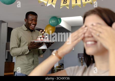 Uomo afroamericano donna sorprendente con torta di compleanno a casa, entrambi sorridenti Foto Stock