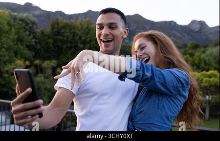 Giovane coppia variegata in giardino che scatta selfie, celebrando il fidanzamento con espressioni gioiose Foto Stock