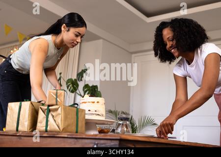 Diverse amiche che preparano torte e regali per festeggiare a casa, sorridendo e divertendosi Foto Stock