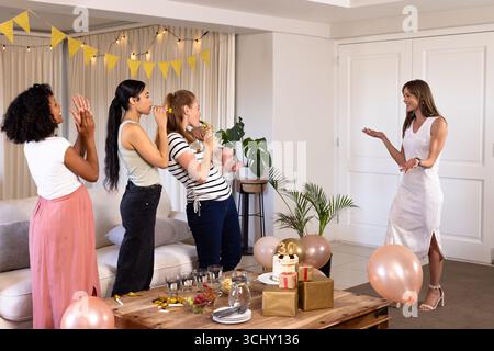 Diverse amiche donne sorprendono la donna con festeggiamenti di compleanno a casa, applaudendo e sorridendo Foto Stock