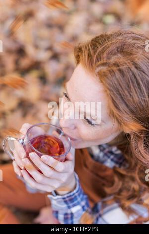 donna dai capelli rossi seduta in foglie autunnali che beve tè caldo Foto Stock