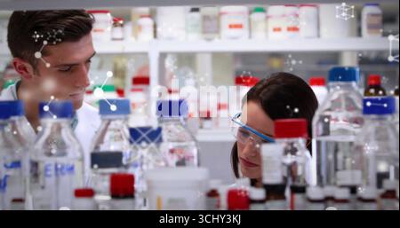 Scienziati di sesso maschile e femminile in camici da laboratorio che manipolano flaconi di reagente da banco, con grafica molecolare Foto Stock
